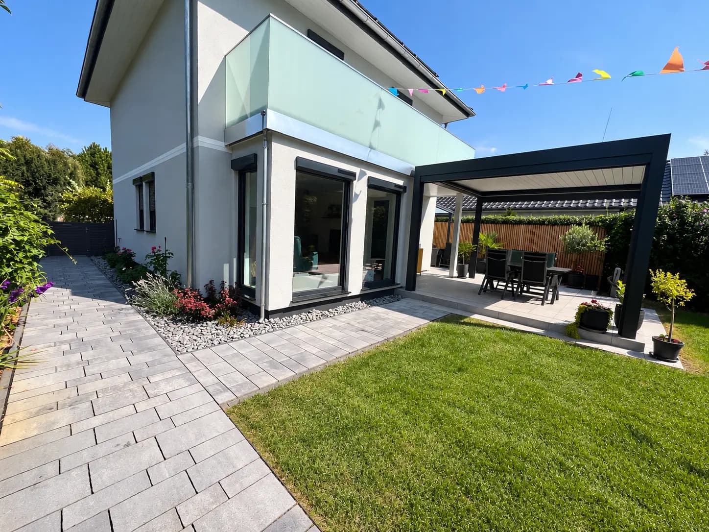 Gepflasterter Weg entlang eines weißen Wohnhauses, dahinter eine überdachte Terrasse mit Sitzgruppe und eine Rasenfläche.