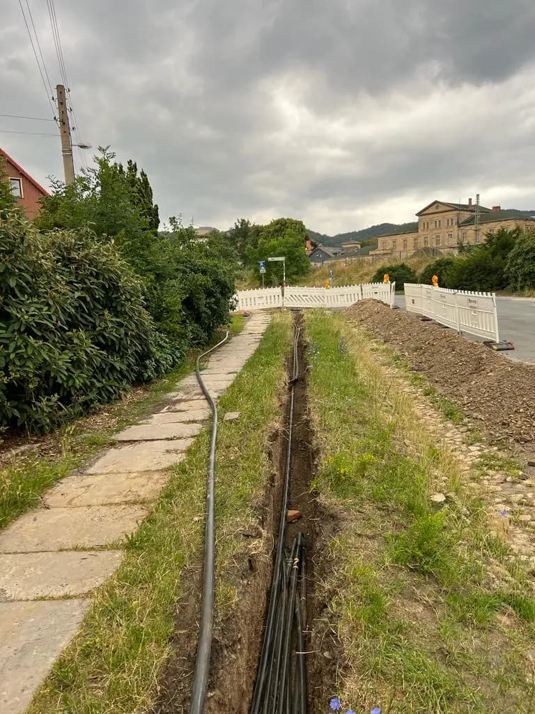 Schmaler Leitungsgraben mit schwarzen Schutzrohren zwischen Plattenweg und Aushub, im Hintergrund eine Absperrung.