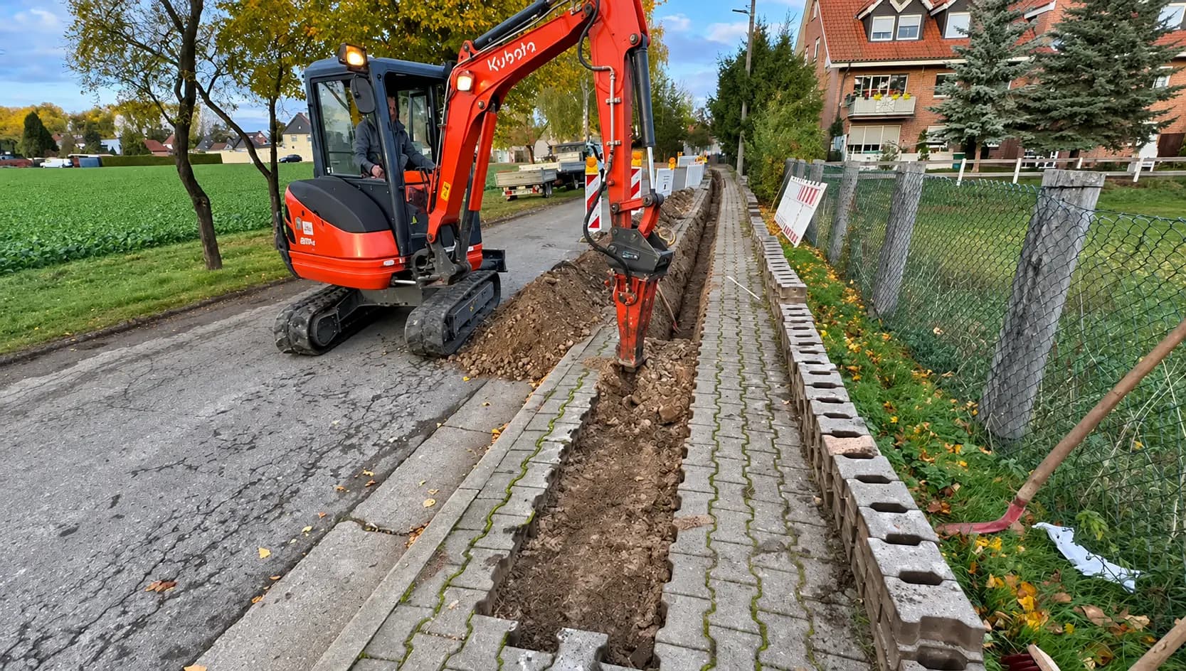 Roter Minibagger hebt entlang eines gepflasterten Gehwegs einen schmalen Leitungsgraben aus, daneben Straße und Zaun.