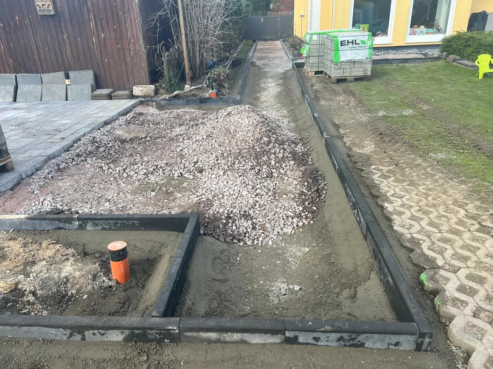 Vorbereitete Wegefläche mit Einfassungen und Materialhaufen zwischen dem gelben Haus und einem Holzschuppen.